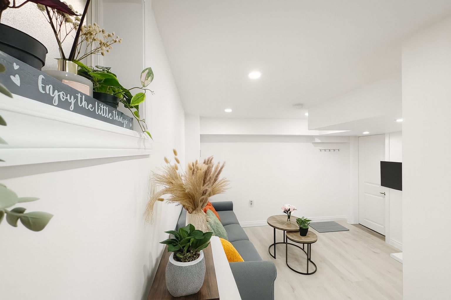 A modern living room with a grey sofa, a small round table, and a decorative wall shelf with plants and a sign.