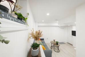 Un salon moderne avec un canapé gris, une petite table ronde et une étagère murale décorative agrémentée de plantes et d’un panneau.