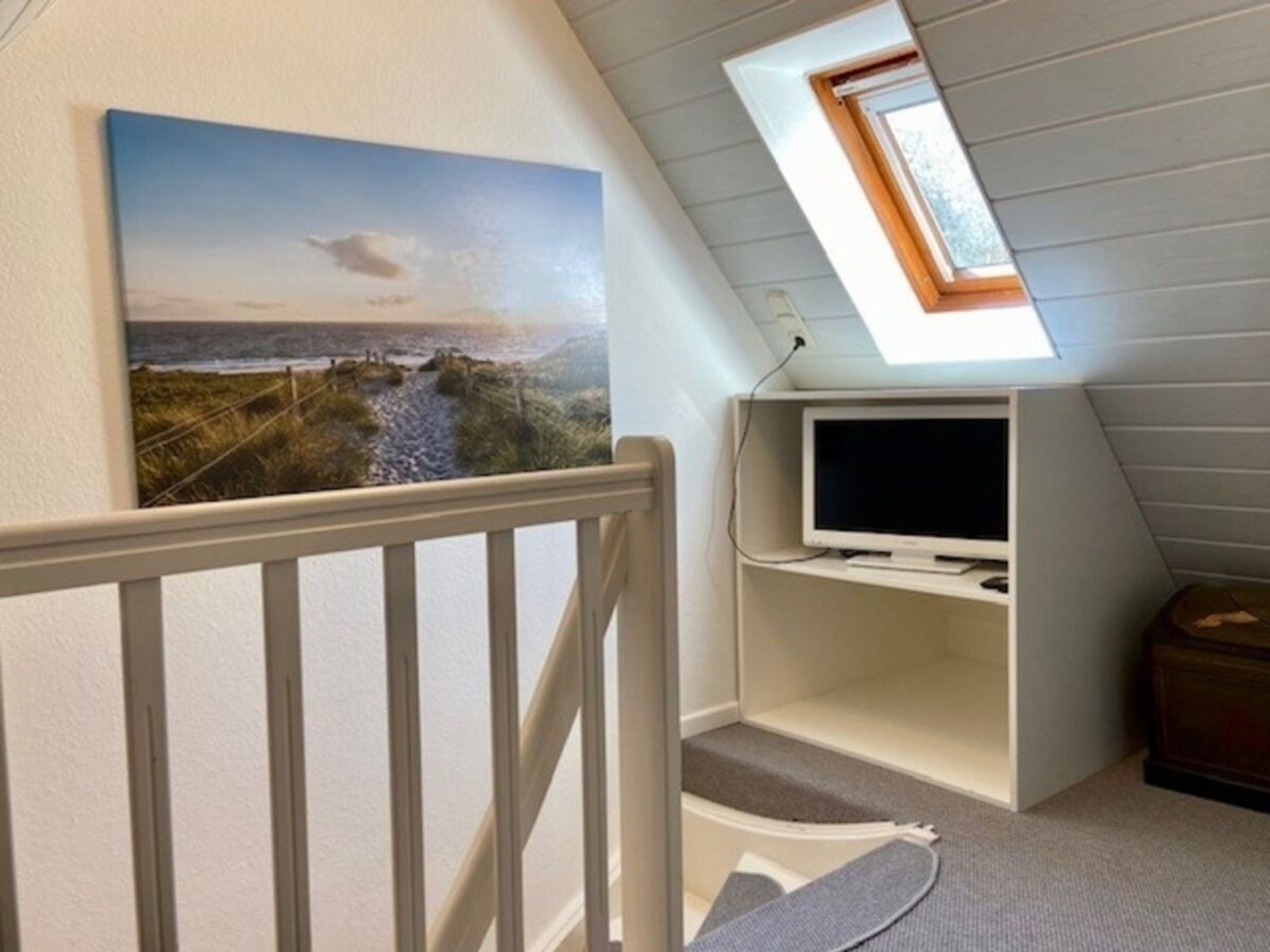 Une pièce avec un plafond en pente, un tableau représentant un paysage côtier, un téléviseur posé sur une étagère et un petit coffre en bois.