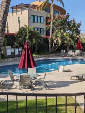 A pool area with lounge chairs and umbrellas, surrounded by palm trees and a building with a curved facade. - Ocean and Golf Views | Walk to Beach & Downtown (San Jose Del Cabo)