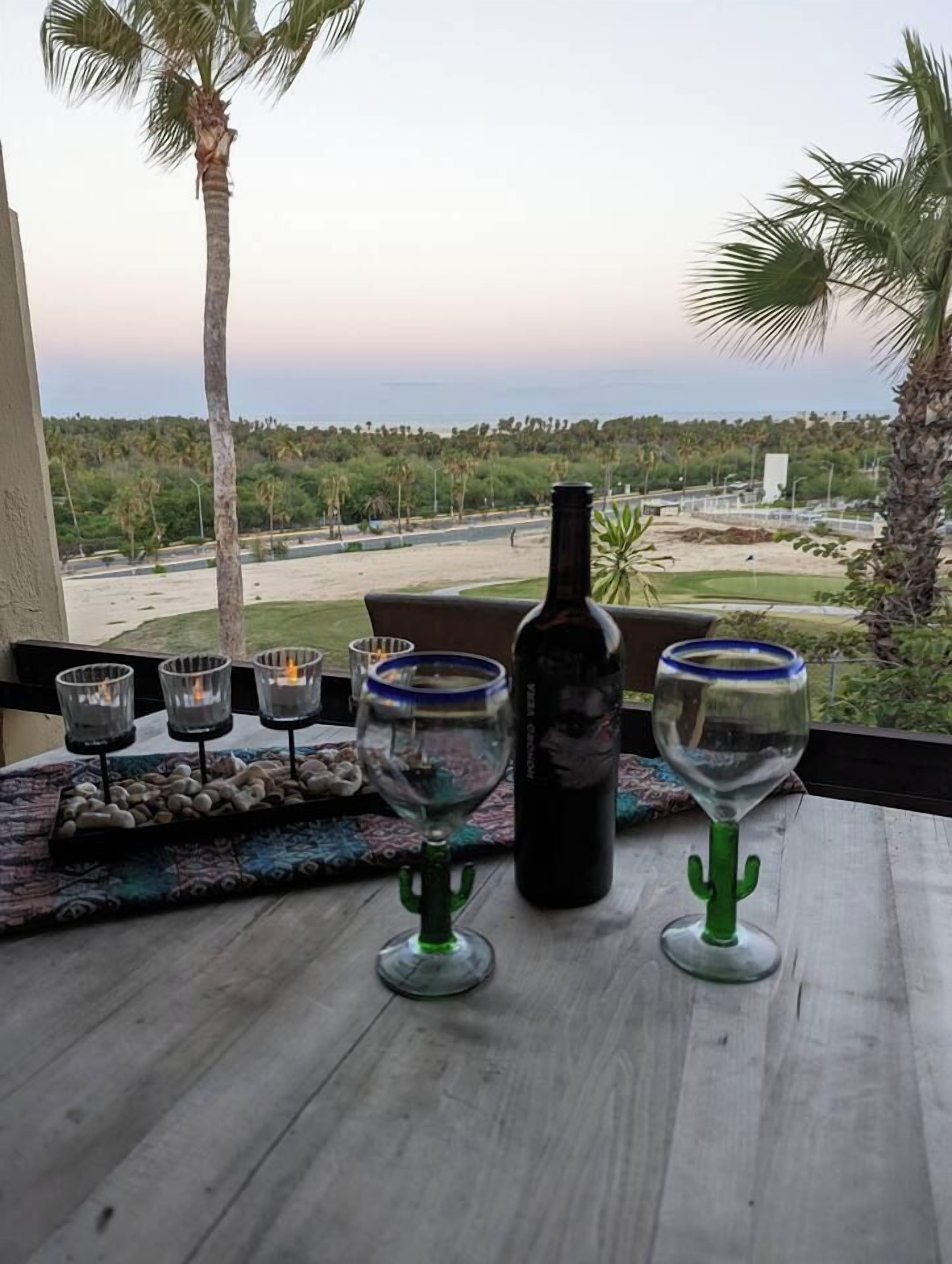 Een tafel met een fles en glazen, met uitzicht op een landschap met palmbomen en een verre horizon.