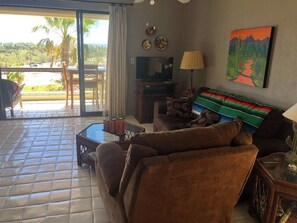 A living room with a sofa, a coffee table, and a TV. There is a painting on the wall and a view of palm trees outside. - Ocean and Golf Views | Walk to Beach & Downtown (San Jose Del Cabo)