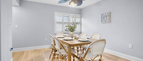 A dining area with a table set for four, surrounded by wicker chairs, a ceiling fan, and a window with blinds.