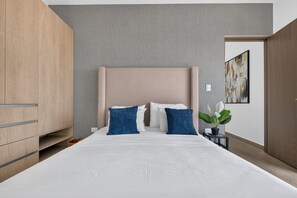 A modern bedroom with a large bed, a wooden wardrobe, and a small bedside table with a plant. - Distrito Corazón: Bright 2BR w/ Balcony & Pool (Playa del Carmen)