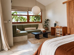 Ein modernes Schlafzimmer mit einem grünen Sofa, einem Holznachttisch, einem Beistelltisch und einem großen Fenster mit Blick ins Grüne.