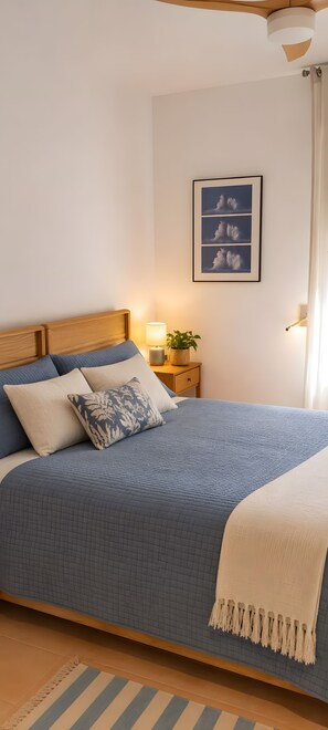 Een netjes opgemaakt bed met een blauwe spreideken en een witte plaid, een nachtkastje met een lamp en een plant, en een lijst met een foto van wolken aan de muur.