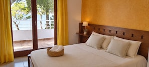 Ein Hotelzimmer mit einem Bett, einem Nachttisch mit Lampe, Blick ins Freie durch einen Balkon und einem mit Vorhängen versehenen Fenster.