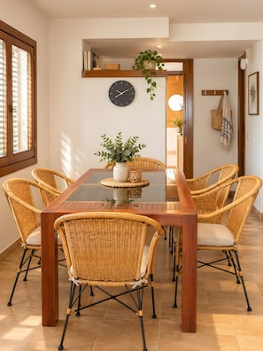 A dining area with a wooden table and wicker chairs, a wall clock, and a potted plant.