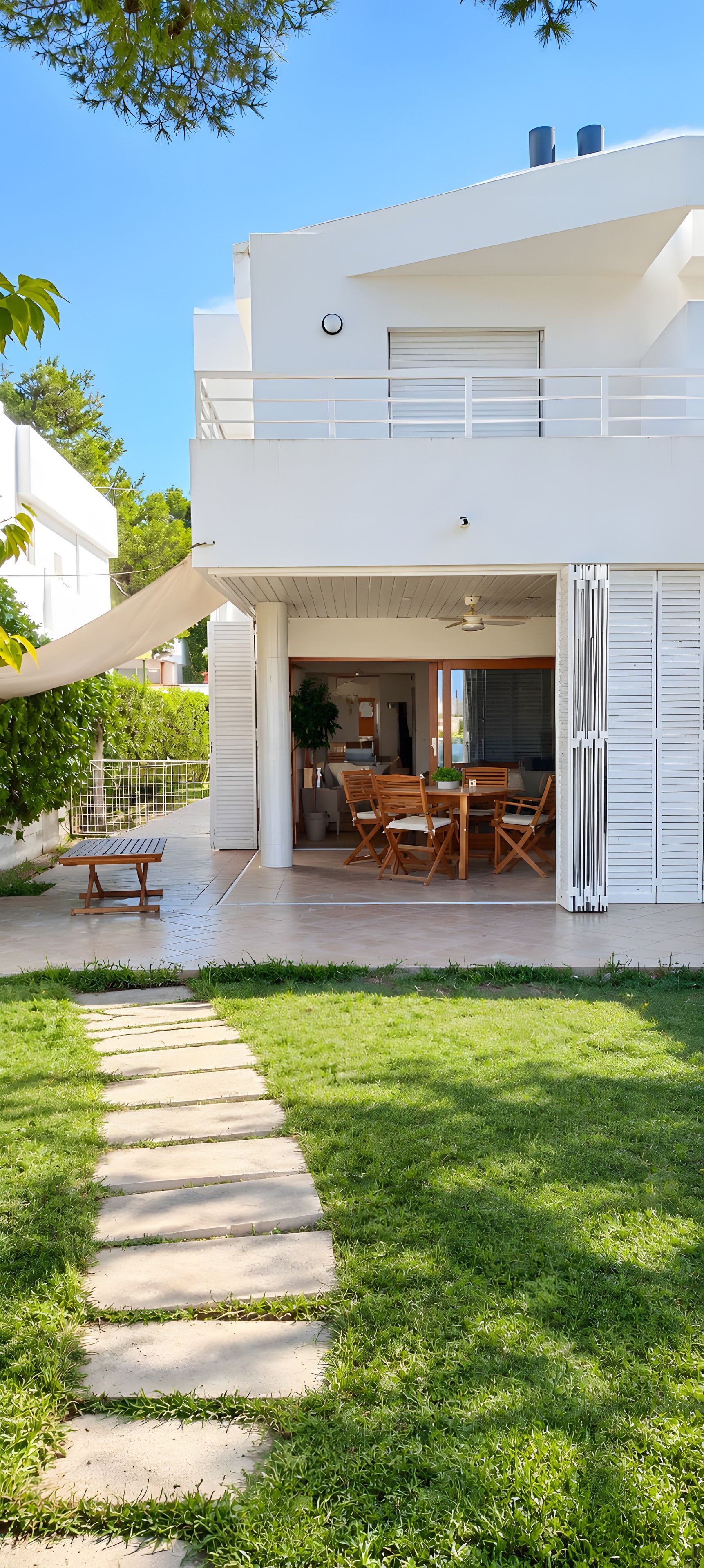 Et moderne hus i to etager med en overdækket terrasse, et spiseområde med træmøbler og en sti til indgangen.