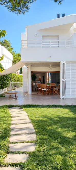 Ein modernes, zweistöckiges Haus mit einer überdachten Terrasse, einem Essbereich mit Holzmöbeln und einem Weg, der zur Eingangstür führt.
