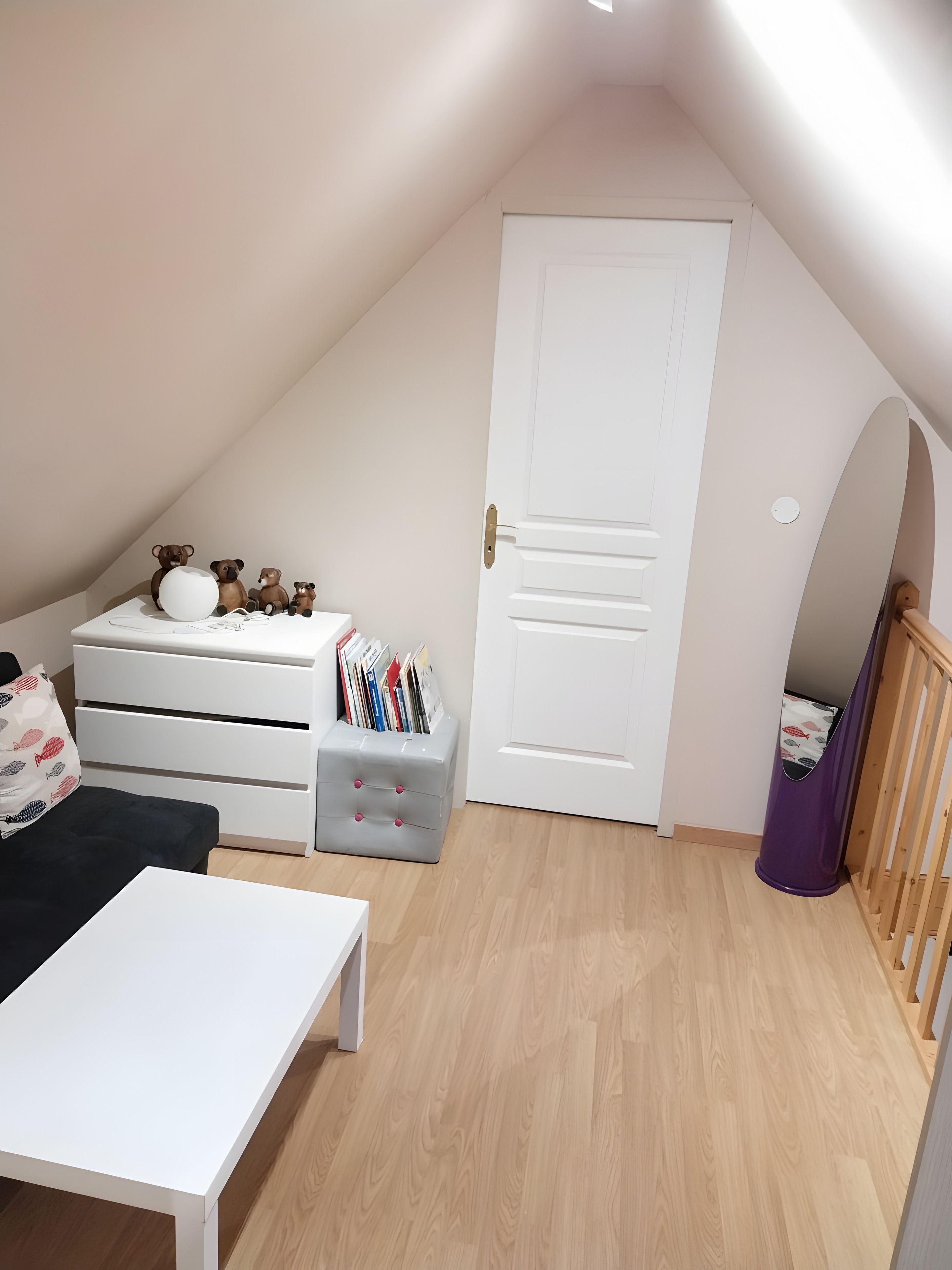 A room with a sloped ceiling, a white door, a bed with a black bedspread, a white table, a grey storage unit with books, and a purple trash bin.
