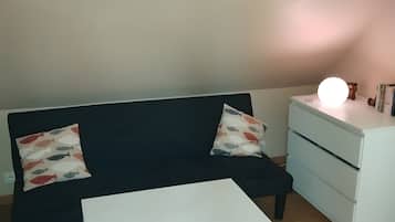 A room with a black sofa, two patterned pillows, a white bedside table, a white nightstand, and a small lamp.