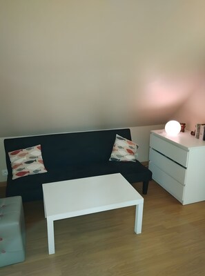 A room with a black sofa, two patterned pillows, a white bedside table, a white nightstand, and a small lamp.