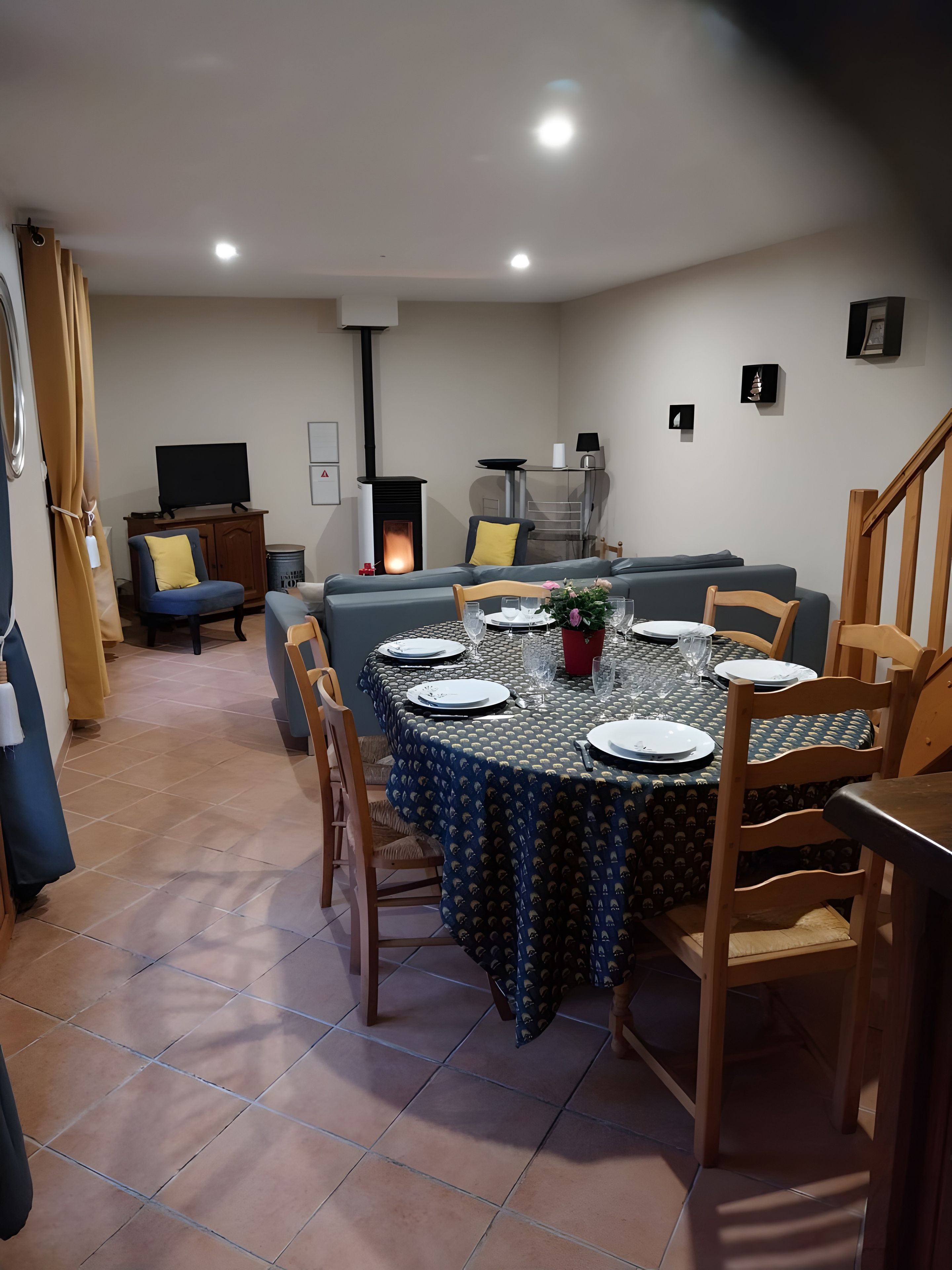 A dining area with a round table set for four, surrounded by chairs. A sofa and a television are visible in the background.