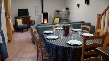 A dining area with a round table set for four, surrounded by chairs. A sofa and a television are visible in the background.