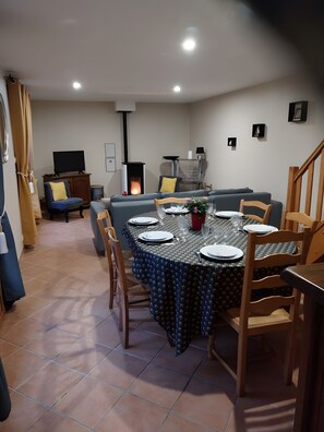 Un comedor con una mesa redonda dispuesta para cuatro personas, rodeada de sillas. Se ve un sofá y un televisor al fondo.