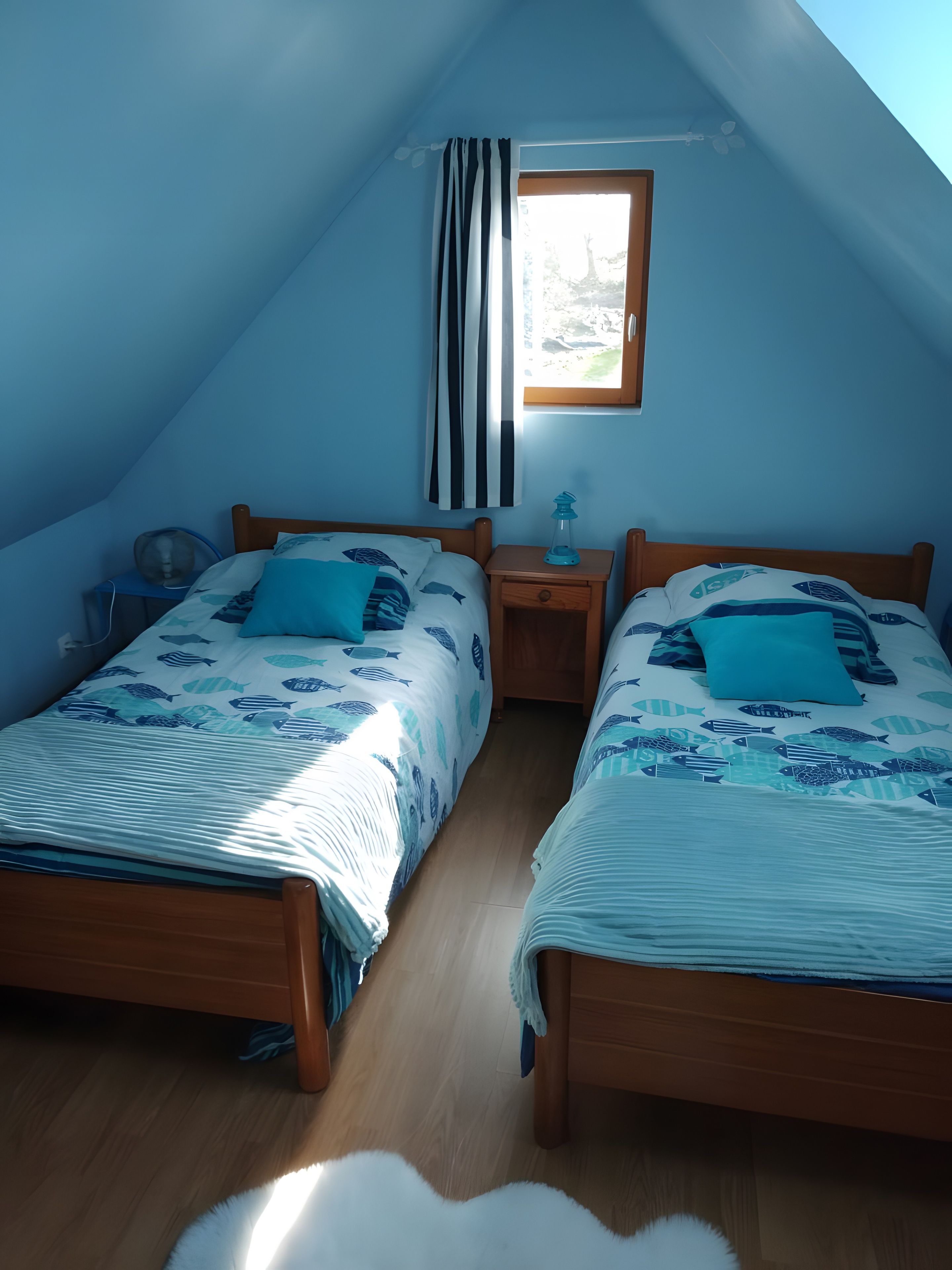 Ein Zimmer mit zwei Betten, einem Fenster mit Vorhängen, einem Nachttisch mit Lampe und einem Deckenventilator.