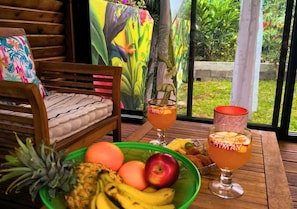 Una terraza de madera con una mesa de comedor, dos bebidas con la etiqueta 'CHIMAY', un cuenco de fruta y una silla de madera con cojín.