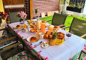 Une table de jardin dressée pour le petit-déjeuner, avec des viennoiseries, des fruits et des boissons.