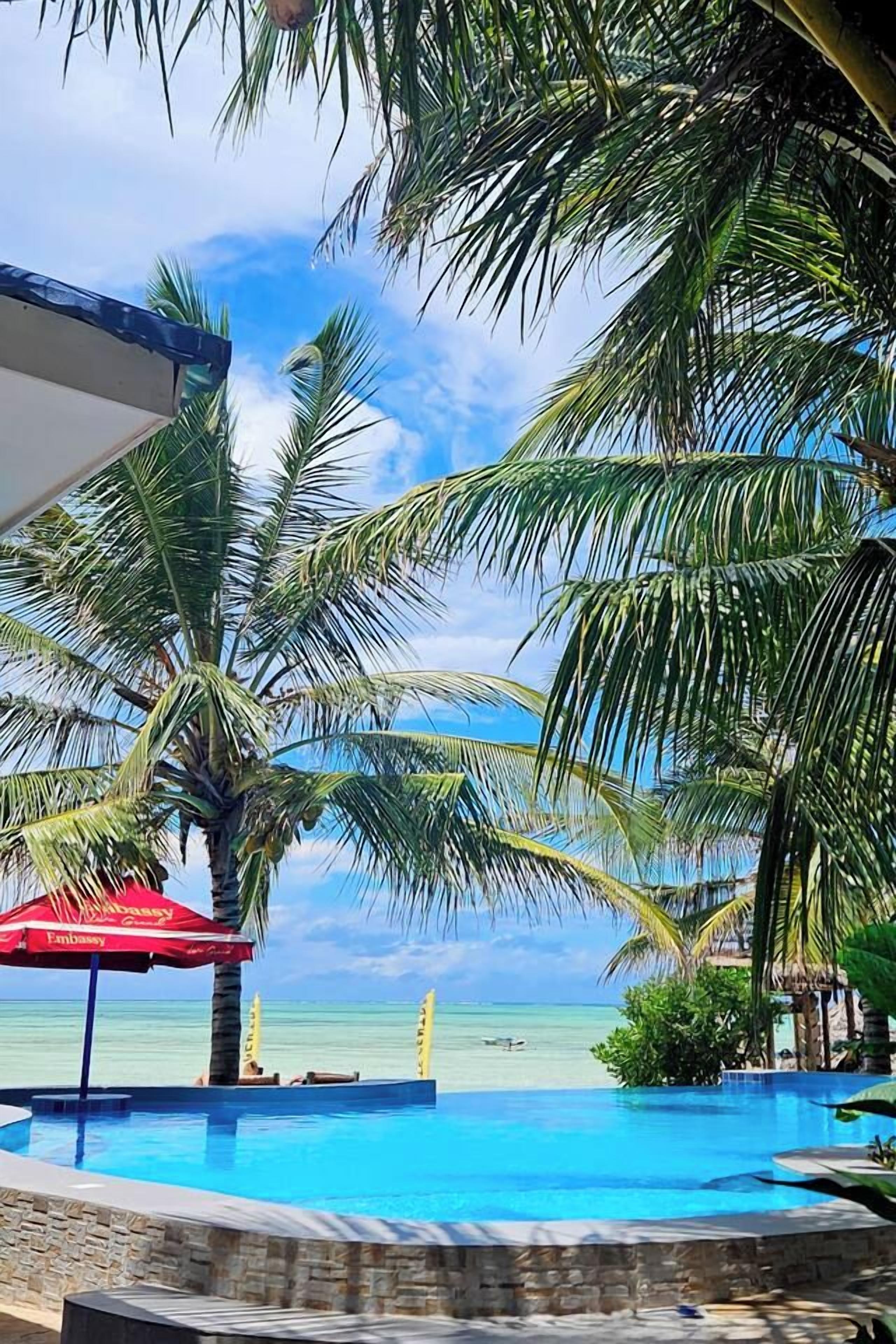 Un espace piscine avec vue sur l’océan, des palmiers et un parasol rouge.