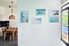 A dining area with a table and chairs, a kitchen with a black countertop, and a wall with framed artwork depicting coastal scenes. - Flat "Zanzi Dream" at The Soul with Pool, balcony, AC (Paje)