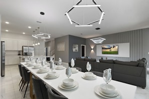 Une cuisine moderne avec des meubles blancs, un coin repas doté d’une longue table dressée pour six personnes, et un canapé gris dans l’espace salon.