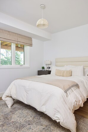 A bedroom with a large bed, a bedside table with a lamp, a window with blinds, and a hanging light fixture. - Brand New LakeHouse StrattCottage (Douro-Dummer)