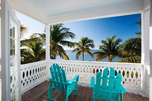 Eine Veranda mit zwei türkisfarbenen Stühlen, weißen Ziergittern und Blick auf Palmen und das Meer.
