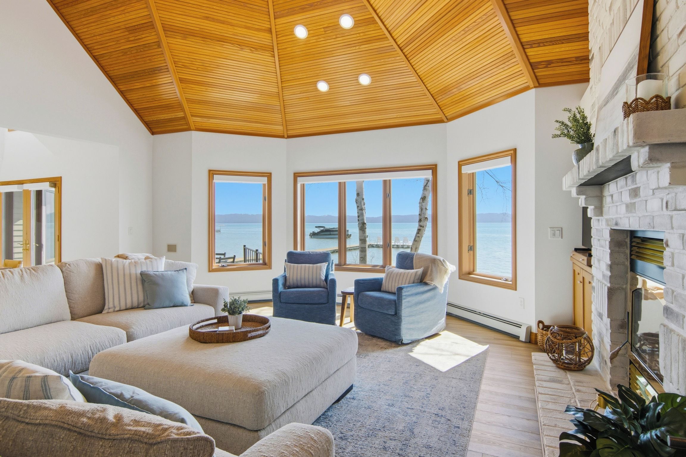A spacious living room with a high wooden ceiling, large windows offering a view of the sea, a fireplace, and a cozy seating area.