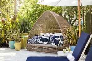 Un fauteuil suspendu en osier, agrémenté de coussins, entouré de plantes en pot et surmonté d’un parasol blanc.