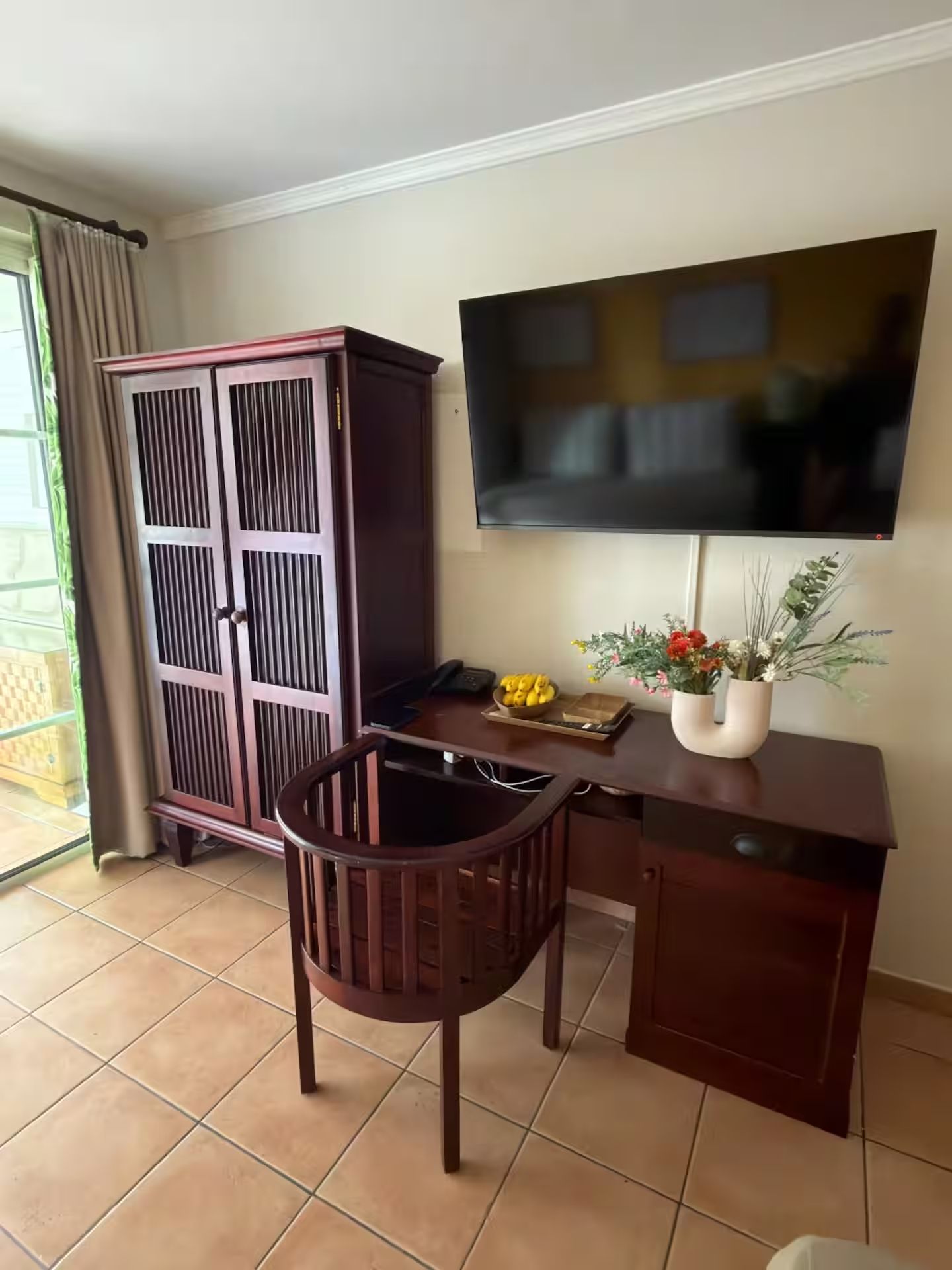 Una habitación con escritorio, silla, televisor, armario y ventana con cortinas.