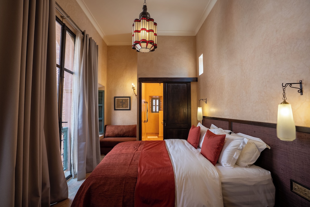 A hotel room with a large bed, a chandelier, a door leading to a bathroom, and a window with curtains.