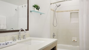 Un baño con un lavamanos blanco, dos llaves, un espejo, un estante de vidrio con una planta, una ducha con cabezal de ducha manual y una bañera.