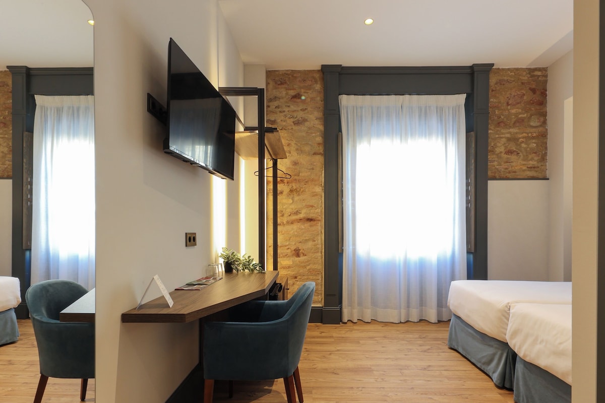 Habitación de hotel con suelo de madera, un escritorio con sillas, una cama, televisor montado en la pared y un espejo grande.