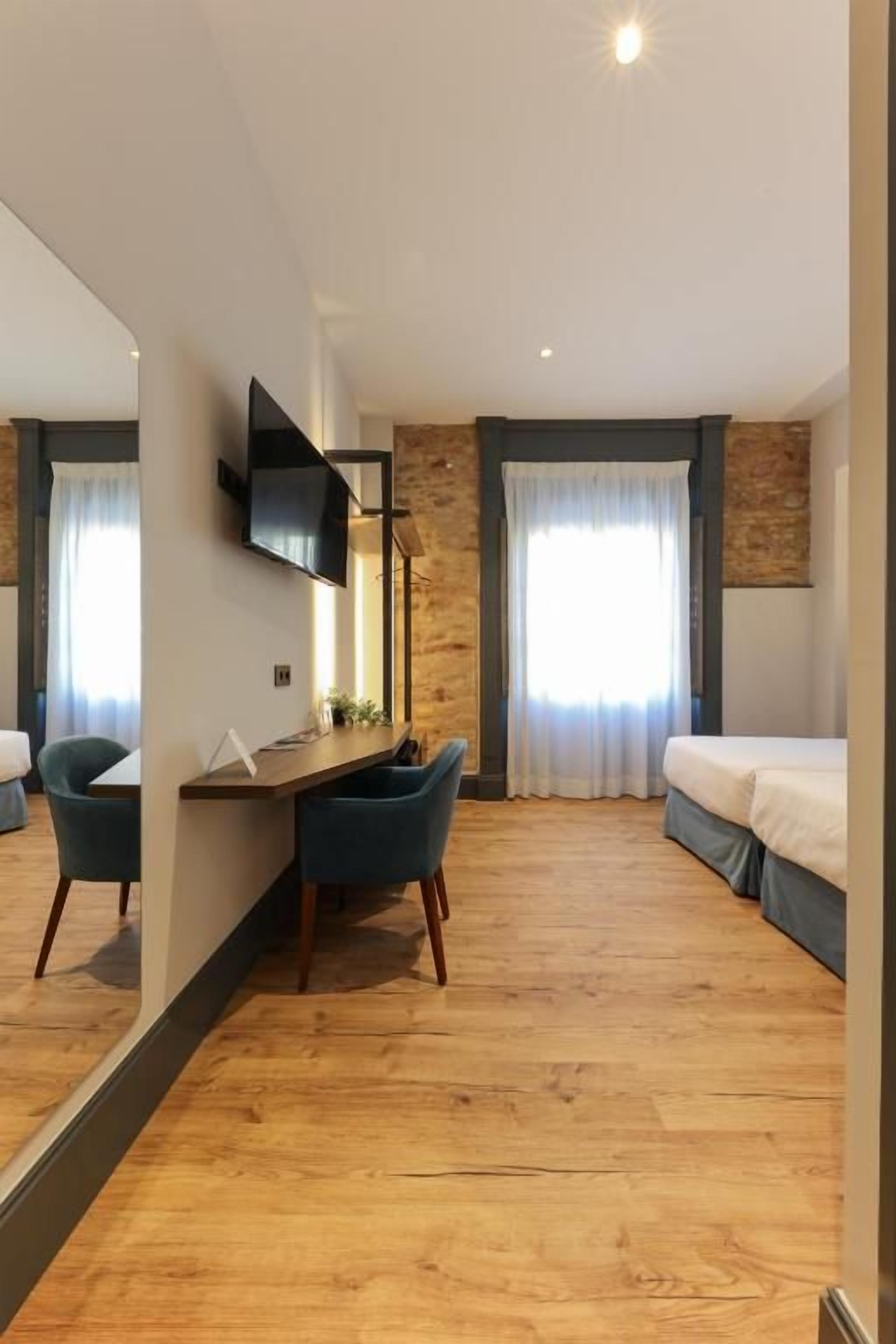 A hotel room with a wooden floor, a desk with chairs, a bed, a TV mounted on the wall, and a large mirror.