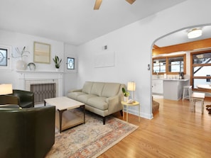 A modern living room with a sofa, coffee table, and chairs, a fireplace, and a kitchen area visible through an archway. - A Stone's Throw | Steps to Lake Michigan (Gary)