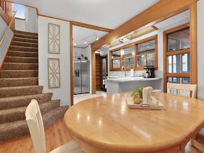 A dining area with a wooden table and chairs, a kitchen in the background, and a staircase leading to another level.
