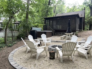 Ein Sitzbereich im Freien mit Adirondack-Stühlen, die um eine Feuerstelle, einen Grill und eine überdachte Konstruktion in einer bewaldeten Umgebung angeordnet sind.