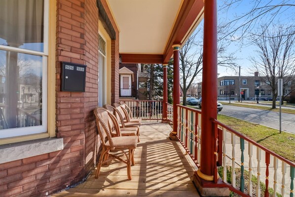 A covered porch with wicker chairs, a brick building, and a street view. - Blarney Bungalow | Renovated Downtown Charmer (Michigan City)