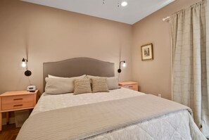 A bedroom with a large bed, two bedside tables, a nightstand, a wall-mounted light, and a curtain. - Blarney Bungalow | Renovated Downtown Charmer (Michigan City)
