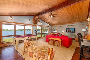 Ein geräumiges Wohnzimmer mit Billardtisch, rotem Sofa und großen Fenstern mit Blick auf ein Gewässer.