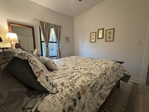 A bedroom with a bed, a nightstand, a lamp, and a window with curtains. - Dragonfly in the Pines | Remodeled Forest Getaway (Michigan City)
