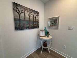 A hallway with a painting of trees, a small table with a framed picture, and a decorative bird. - Dragonfly in the Pines | Remodeled Forest Getaway (Michigan City)