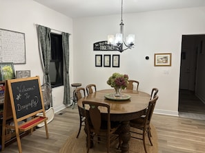A dining area with a round wooden table, chairs, a chandelier, and a chalkboard sign. - Dragonfly in the Pines | Remodeled Forest Getaway (Michigan City)