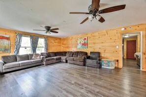 Ein geräumiges Wohnzimmer mit einer Eckcouch, einem einzelnen Sessel und einem Couchtisch. Zwei Deckenventilatoren und ein Fenster mit Vorhängen sind vorhanden.