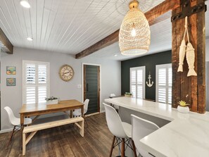 A dining area with a wooden table and white chairs, a hanging light fixture, and a wall clock. - Dune Blast Cottage | Hot Tub, Fire Pit & Beach (Michigan City)