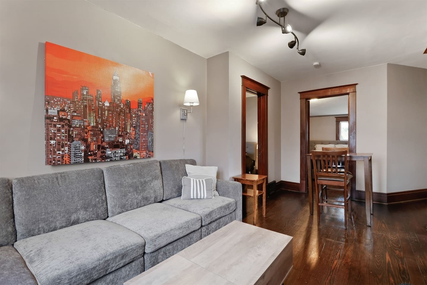 A modern living room with a gray sectional sofa, a coffee table, a cityscape painting, a dining area with a wooden table and chairs, and a hallway with a wooden door.