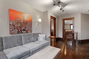 A modern living room with a gray sectional sofa, a coffee table, a cityscape painting, a dining area with a wooden table and chairs, and a hallway with a wooden door. - The Gable at 409 | Craftsman Charm in Downtown (Michigan City)