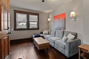 A modern living room with a grey sectional sofa, a wooden coffee table, and a bench. There are two windows with blinds, a cityscape painting on the wall, and two wall-mounted lamps. - The Gable at 409 | Craftsman Charm in Downtown (Michigan City)
