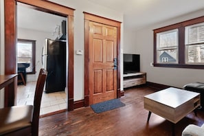A room with a wooden door, a television on a stand, a dining table, and a window with blinds. - The Gable at 409 | Craftsman Charm in Downtown (Michigan City)